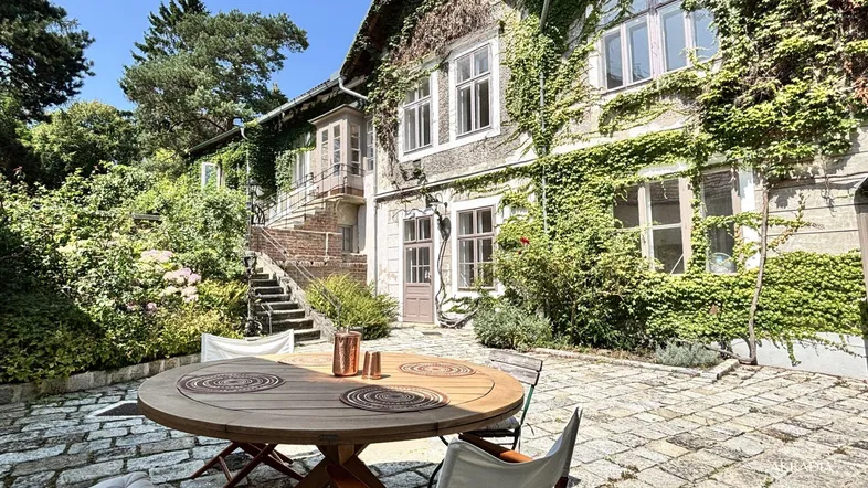Gemütliche Terrasse mit Holztisch und Stühlen, umgeben von üppigem Grün und einem historischen Gebäude.