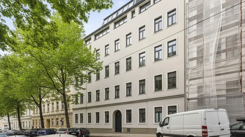 Fassade eines Mehrfamilienhauses an einer Stadtstraße mit parkenden Autos und Bäumen.