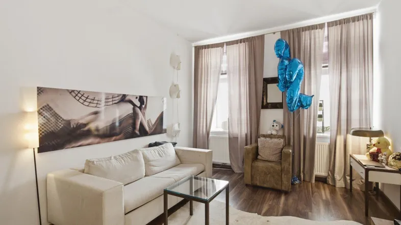 Helles Wohnzimmer mit beigem Sofa, Holzboden und großen Fenstern mit Vorhängen.
