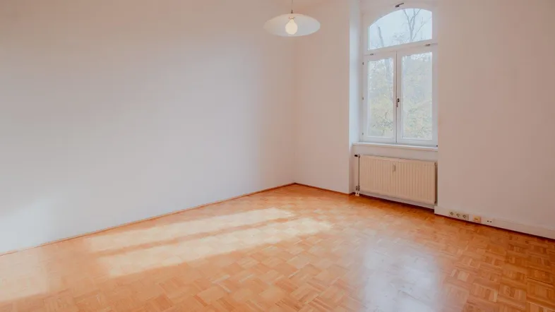 Helles Zimmer mit Fischgrätparkett, hohen Decken und einem großen Bogenfenster.