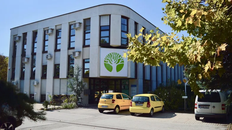Modernes Bürogebäude mit mehreren Etagen, umgeben von Bäumen und Parkplätzen unter blauem Himmel.