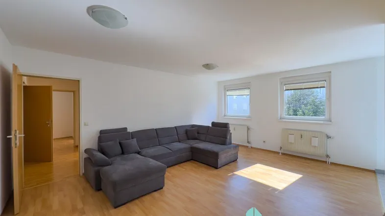 Helles Wohnzimmer mit großem Ecksofa, Parkettboden und zwei Fenstern, die Tageslicht hereinlassen.