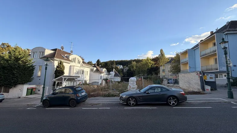 Blick auf eine Straße mit einem unbebauten Grundstück und umliegenden Wohngebäuden unter blauem Himmel.