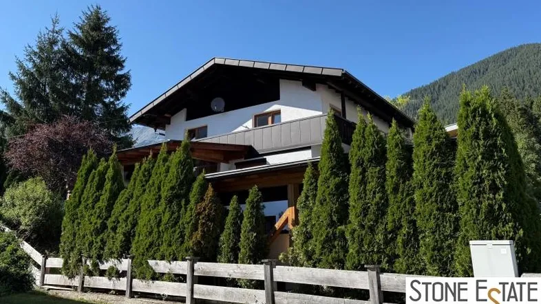 Hausfassade im alpinen Stil mit gepflegter Hecke und Bergblick unter blauem Himmel.