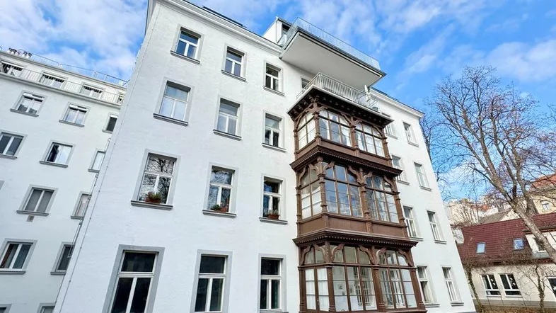 Weißes Altbaugebäude mit traditionellem Holzerker und moderner Dachterrassen-Erweiterung unter blauem Himmel.