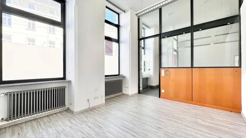 Heller Büroraum mit großen Fenstern, hellem Laminatboden und einer Glaswandabtrennung zu einem weiteren Bereich.