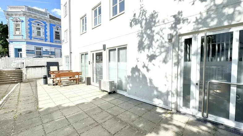 Eine gepflasterte Terrasse mit Holzbänken und Tischen vor einem weißen Gebäude, im Hintergrund ein blaues Haus.