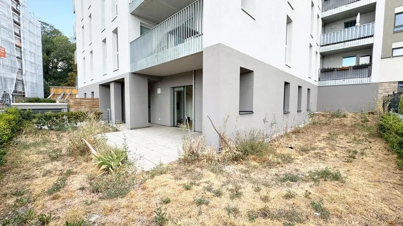 Außenansicht eines Neubaus mit gepflasterter Terrasse und angrenzender Grünfläche im Erdgeschoss.