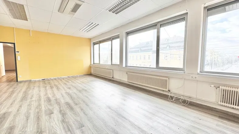 Geräumiger Büroraum mit hellem Laminatboden, gelber Akzentwand und großen Fenstern für viel Tageslicht.