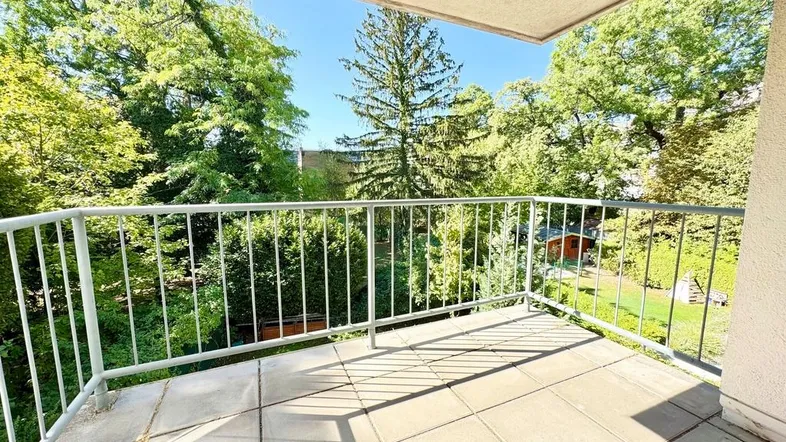 Heller Balkon mit Fliesenboden und Blick ins Grüne, umgeben von hohen Bäumen.