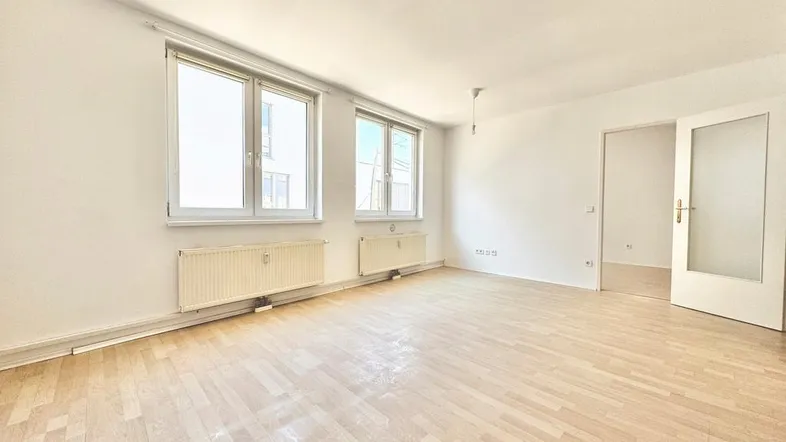 Helles, leeres Wohnzimmer mit zwei großen Fenstern, Parkettboden und weiß gestrichenen Wänden.