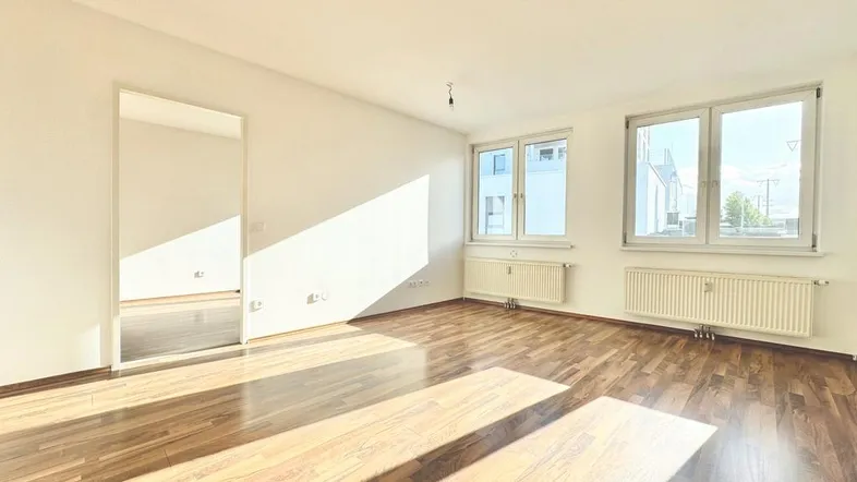 Helles, leeres Wohnzimmer mit Laminatboden und großen Fenstern, durch die viel Sonnenlicht einfällt.