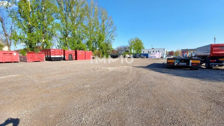 Weitläufiges, unbefestigtes Grundstück mit Lagercontainern und einem LKW im Hintergrund unter blauem Himmel.