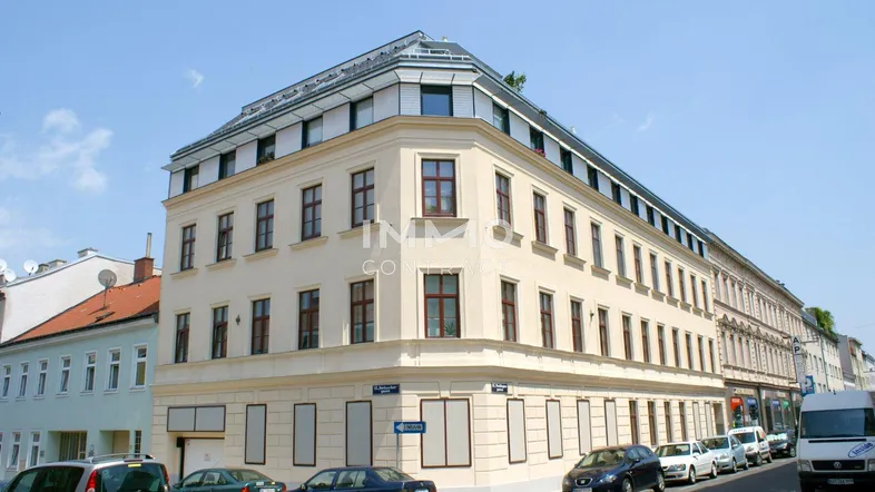 Eckgebäude mit klassischer Fassade und moderner Dachaufstockung unter blauem Himmel.