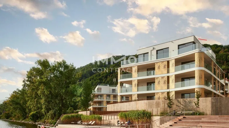 Moderne Wohngebäude am Flussufer mit Terrassen und Bergblick unter bewölktem Himmel.