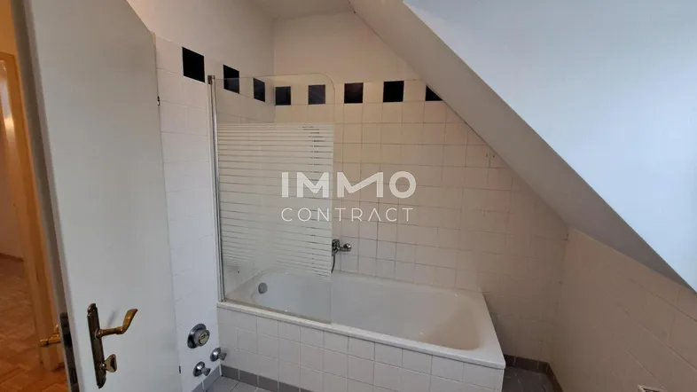 Badezimmer mit Badewanne, Duschwand und Fliesenboden unter einer Dachschräge.