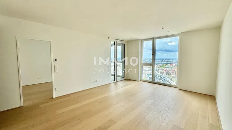 Helles, leeres Wohnzimmer mit Holzboden und großen Fenstern, die einen weiten Stadtblick bieten.