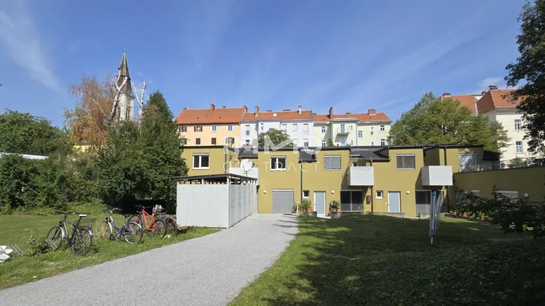 Außenansicht eines Wohngebäudes mit Grünflächen, Kiesweg und Kirchturm im Hintergrund unter blauem Himmel.