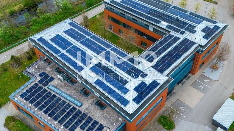Luftaufnahme eines modernen Bürogebäudes mit zahlreichen Solarmodulen auf dem Dach, umgeben von grünen Feldern und einem Fluss.