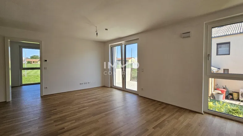 Helles, leeres Wohnzimmer mit Parkettboden und Zugang zu einem Balkon oder einer Terrasse.