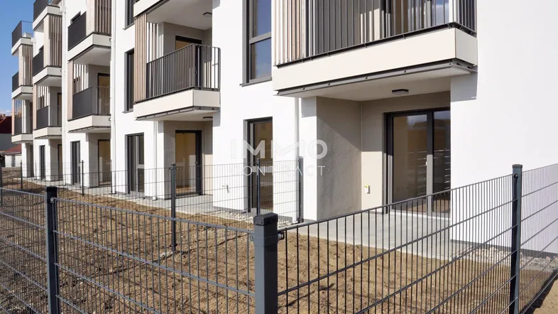 Modernes Neubau-Wohngebäude mit weißen Fassaden, Balkonen und Erdgeschoss-Terrassen, umgeben von einem Zaun.