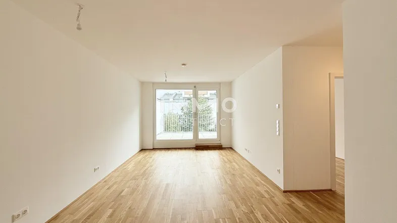 Helles, leeres Wohnzimmer mit Holzboden und Zugang zu einem Balkon.