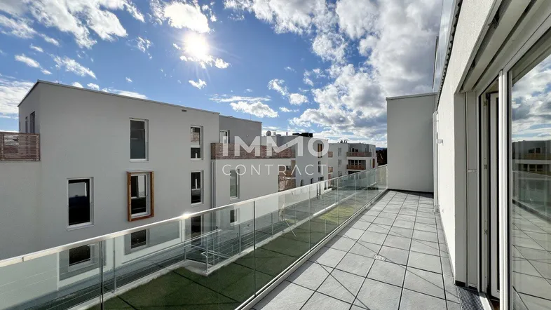 Große, sonnige Terrasse mit modernen Fliesen, Glasgeländer und Blick auf umliegende Neubauten.