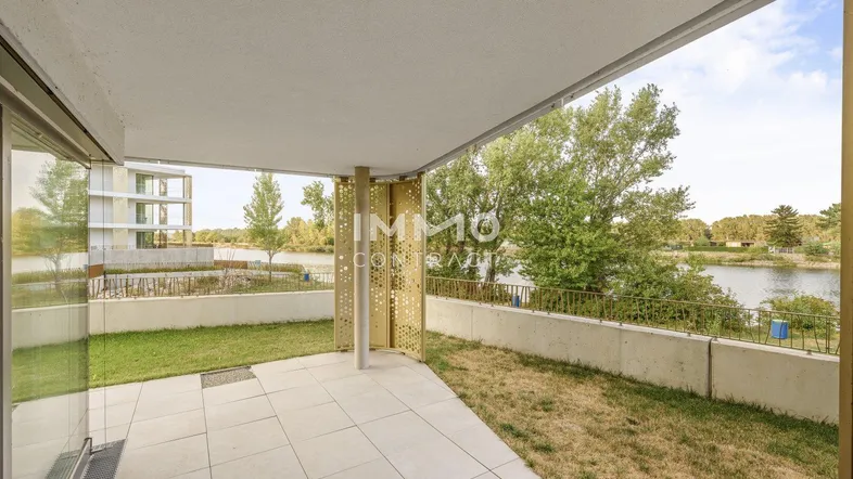 Überdachte Terrasse mit Fliesenboden, Rasenfläche und Blick auf das Wasser, umgeben von moderner Architektur.