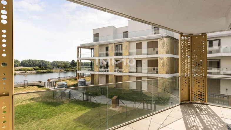 Geräumiger Balkon mit Fliesenboden und Glasgeländer, bietet Blick auf moderne Gebäude und Wasser.