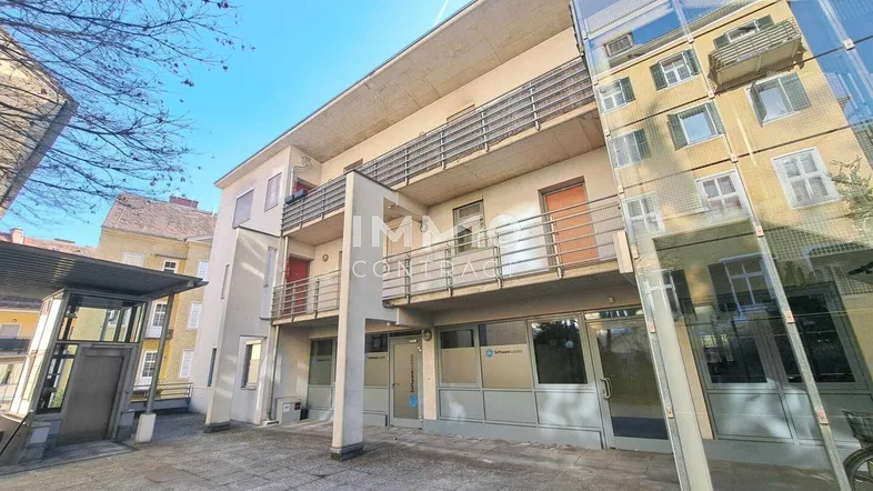 Mehrstöckiges Gebäude mit Balkonen und einem modernen Glasanbau am Eingangsbereich unter blauem Himmel.