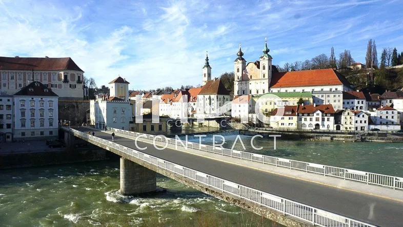 Panoramablick auf eine historische Stadt am Fluss mit Brücke und traditionellen Gebäuden unter blauem Himmel.