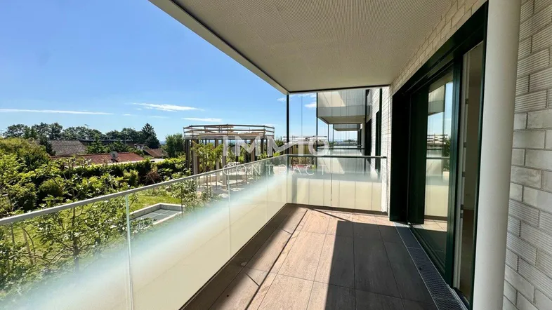 Geräumiger Balkon mit Glasgeländer, Holzboden und weitem Blick ins Grüne unter blauem Himmel.