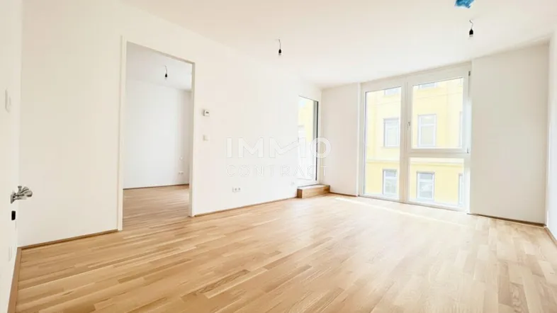 Heller Wohnraum mit großen Fenstern, Parkettboden und Zugang zu weiteren Zimmern.
