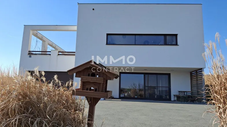 Moderne weiße Villa mit Flachdach und großen Fenstern, umgeben von trockener Vegetation.