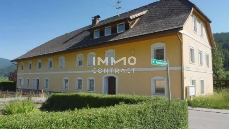 Großes gelbes Gebäude mit traditionellem Satteldach, umgeben von grüner Landschaft und Hecken.