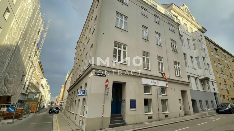 Graues Mehrfamilienhaus an einer Straßenecke in städtischer Umgebung mit Geschäften im Erdgeschoss.