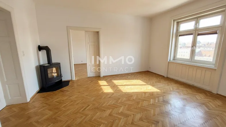 Helles Wohnzimmer mit Fischgrätparkettboden, Kaminofen und Tageslicht durch ein großes Fenster.