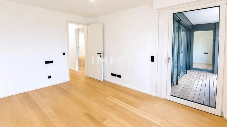 Helles, leeres Zimmer mit Holzboden und Zugang zu einem Balkon oder einer Loggia.