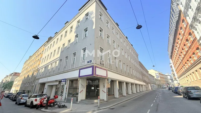 Großes Eckgebäude an einer belebten Straße mit vielen Fenstern und Geschäftsflächen im Erdgeschoss.