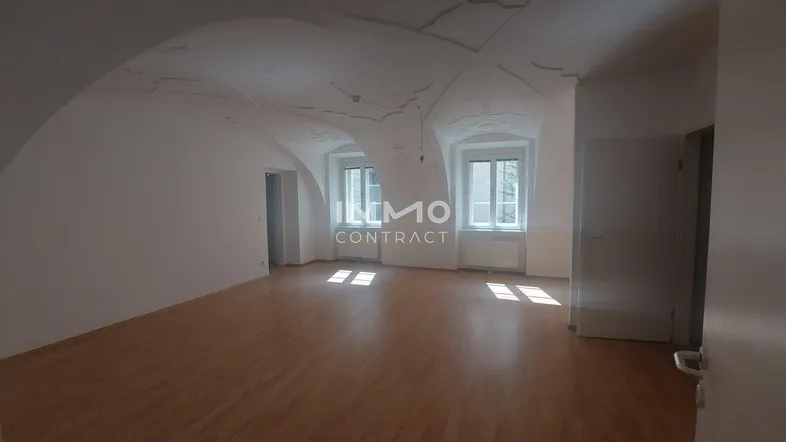 Geräumiges Zimmer mit Holzboden, hohen Fenstern und einer beeindruckenden Gewölbedecke mit Stuckverzierungen.