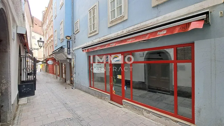 Schmale Gasse mit Kopfsteinpflaster und einer Ladenfront mit großen roten Fenstern und Markise.