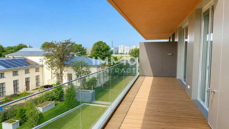 Langer Balkon mit Holzboden, Glasgeländer und Blick auf die grüne Umgebung.