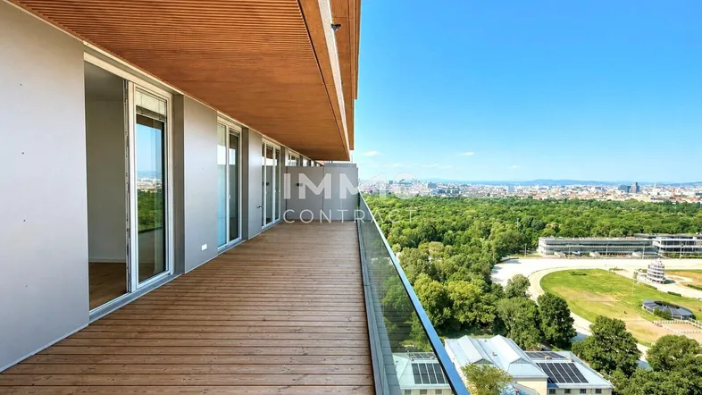 Geräumiger Balkon mit Holzboden und Glasgeländer, bietet weitreichenden Blick über die Stadt und Grünflächen.