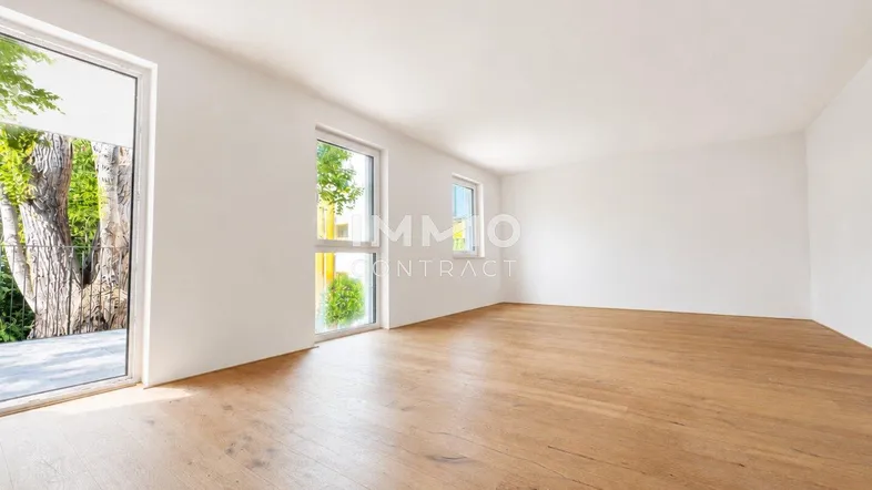 Helles, leeres Zimmer mit Holzboden und Zugang zu einem Außenbereich mit viel Grün.