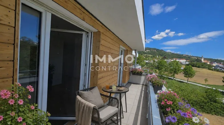 Möblierter Balkon mit Holzwänden, Blumenkästen und weitem Blick auf die Landschaft und den Ort.