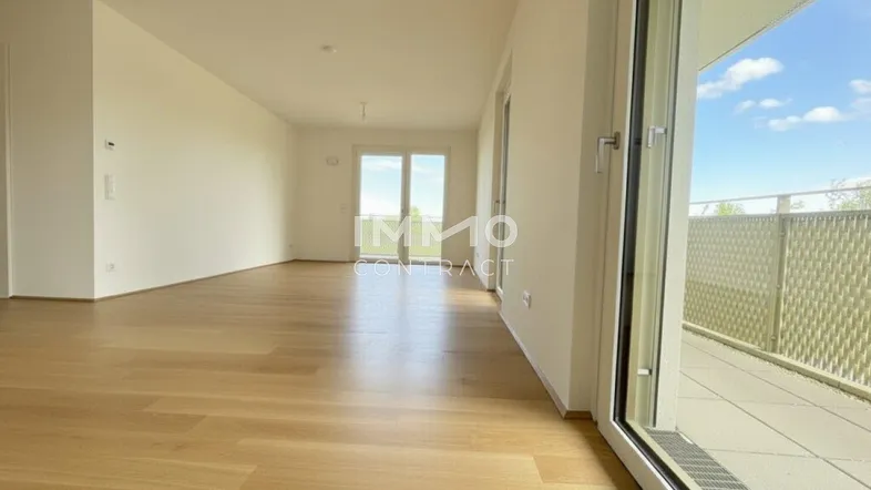 Helles, leeres Wohnzimmer mit Holzboden und Zugang zu einem Balkon.
