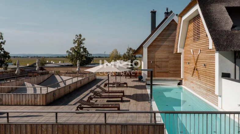 Außenbereich mit Pool, Holzdeck und Liegestühlen, umgeben von modernen Gebäuden mit Reetdächern.