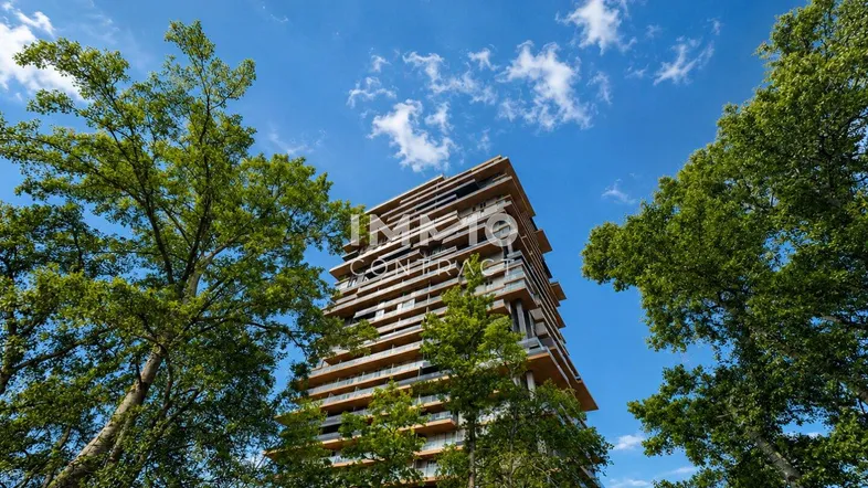 Modernes Hochhaus mit vielen Balkonen, umgeben von Bäumen unter blauem Himmel.