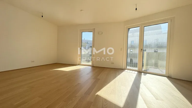 Helles, leeres Zimmer mit Holzboden und Zugang zu einem Balkon, durchflutet von natürlichem Sonnenlicht.