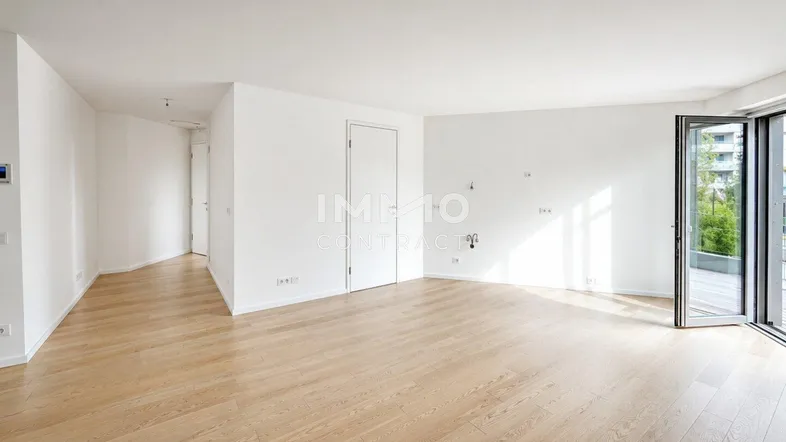 Helles, leeres Zimmer mit Holzboden und Zugang zu einem Balkon oder einer Terrasse.